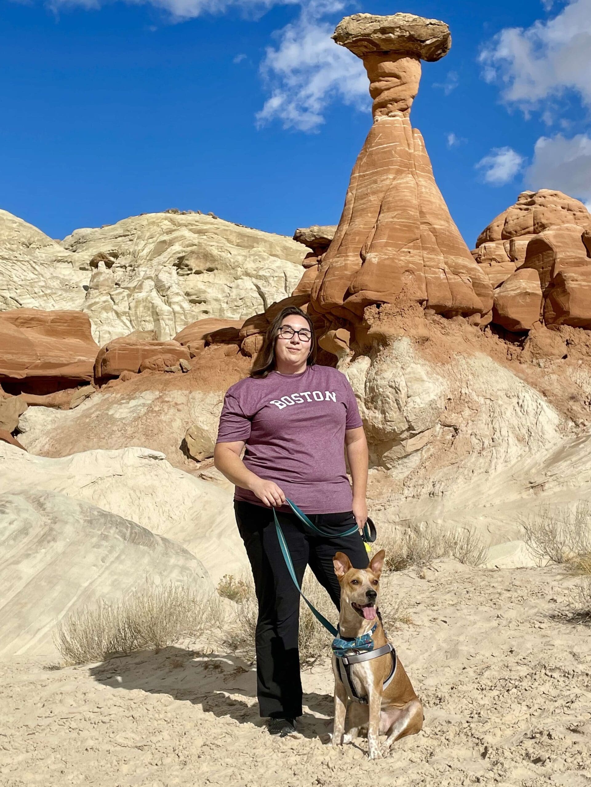 Woman standing with a dog on a leash in a desert rock landscape beneath a tall hoodoo rock formation and bright blue sky with scattered clouds.