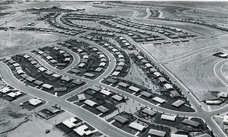 Black-and-white aerial photograph of a suburban neighborhood with curved streets and rows of small single-story homes arranged in planned housing developments.