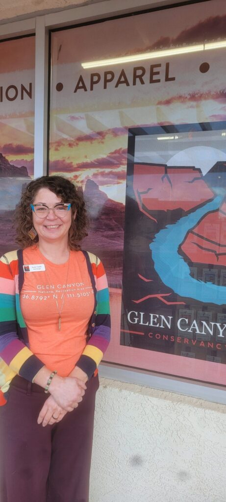 Smiling Glen Canyon Conservancy staff member standing outside an apparel section display wearing a colorful striped cardigan and Glen Canyon National Recreation Area shirt.